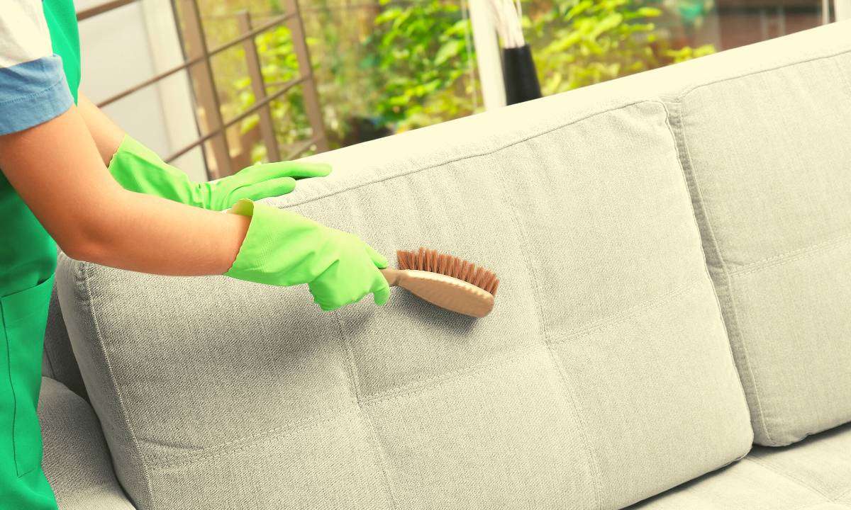 A woman wearing green gloves while brushing a white couch.