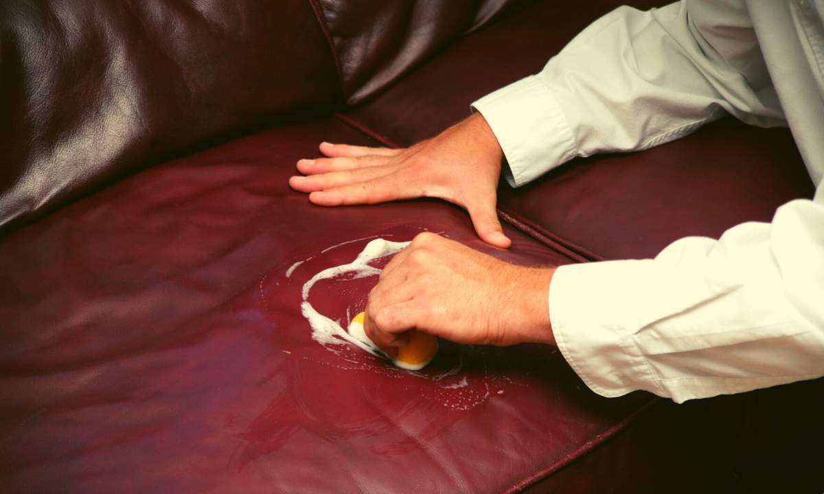A man cleans a dark red leather couch with soap and water.