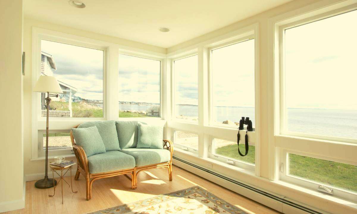 A loveseat with pale blue-green sofa cushions is in the corner of a room with floor-to-ceiling big windows looking out onto a beach and the ocean.