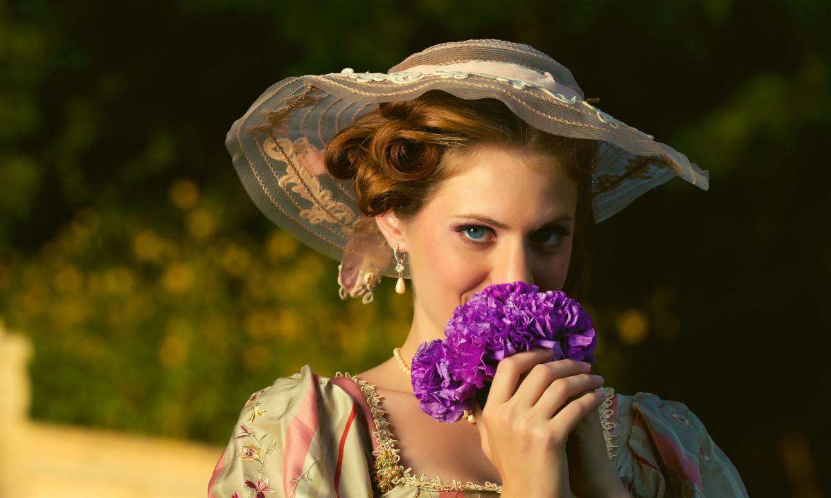 A Victorian woman wearing a large hat and holding purple flowers in front of her face.