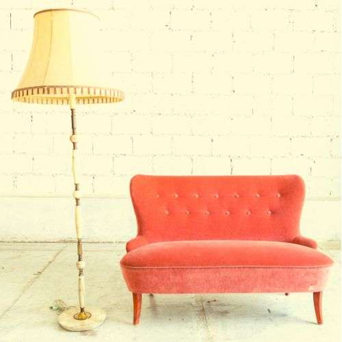 A small, orange-red couch with a lamp next to it against a white wall.
